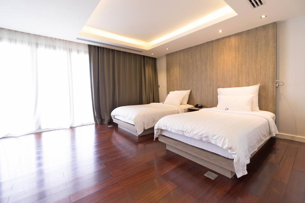 A modern hotel room with two beds, wooden flooring, a large window with curtains, and a wooden accent wall.