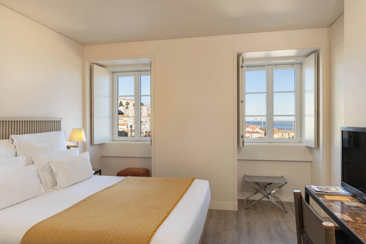 A hotel room with a bed, a desk, a TV, and two windows offering a view of the sea.
