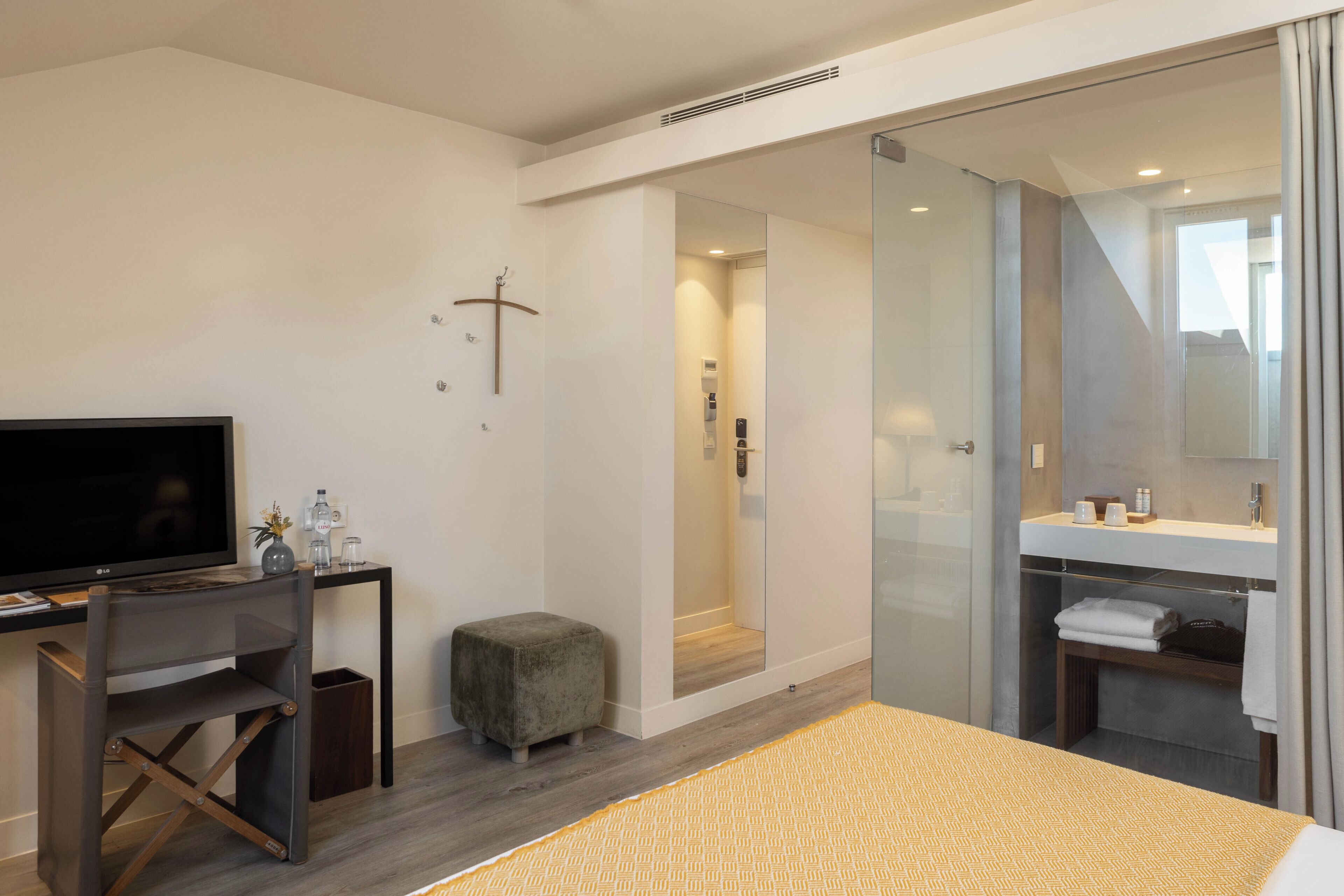 A modern hotel room with a bed, a desk with a flat-screen TV, a small stool, a glass shower enclosure, and a bathroom with a mirror and towel rack.