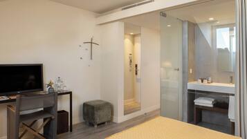 A modern hotel room with a bed, a desk with a flat-screen TV, a small stool, a glass shower enclosure, and a bathroom with a mirror and towel rack.