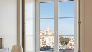 A hotel room with a bed, a window with a view of a cityscape, and a small orange ottoman. - Memmo Alfama - Design Hotels (Lisbon)