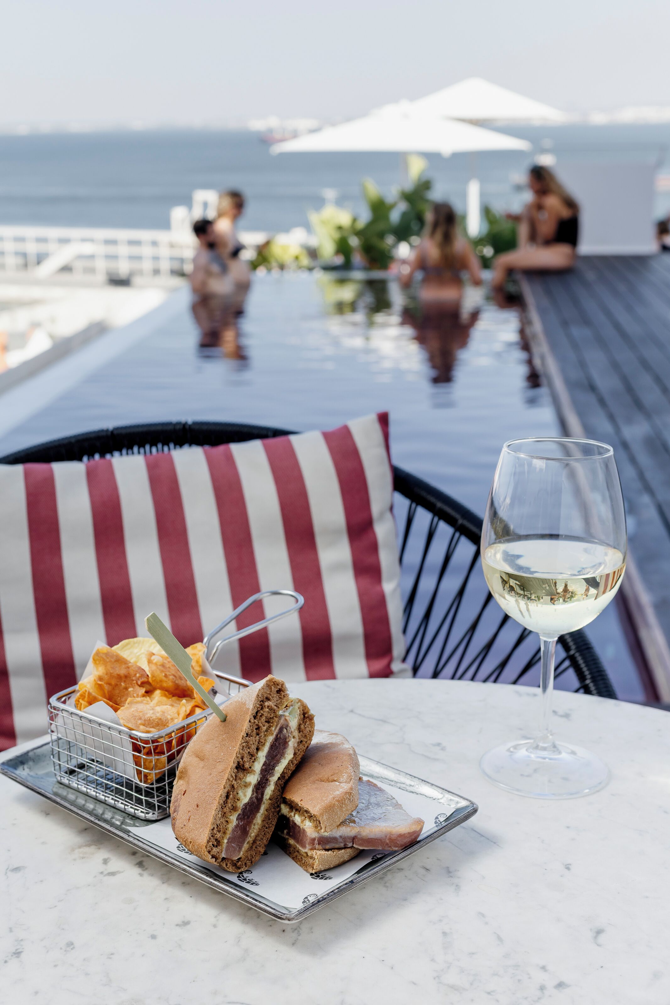 A dining table with a sandwich, chips, and a glass of white wine, overlooking a pool area with people and a view of the sea.