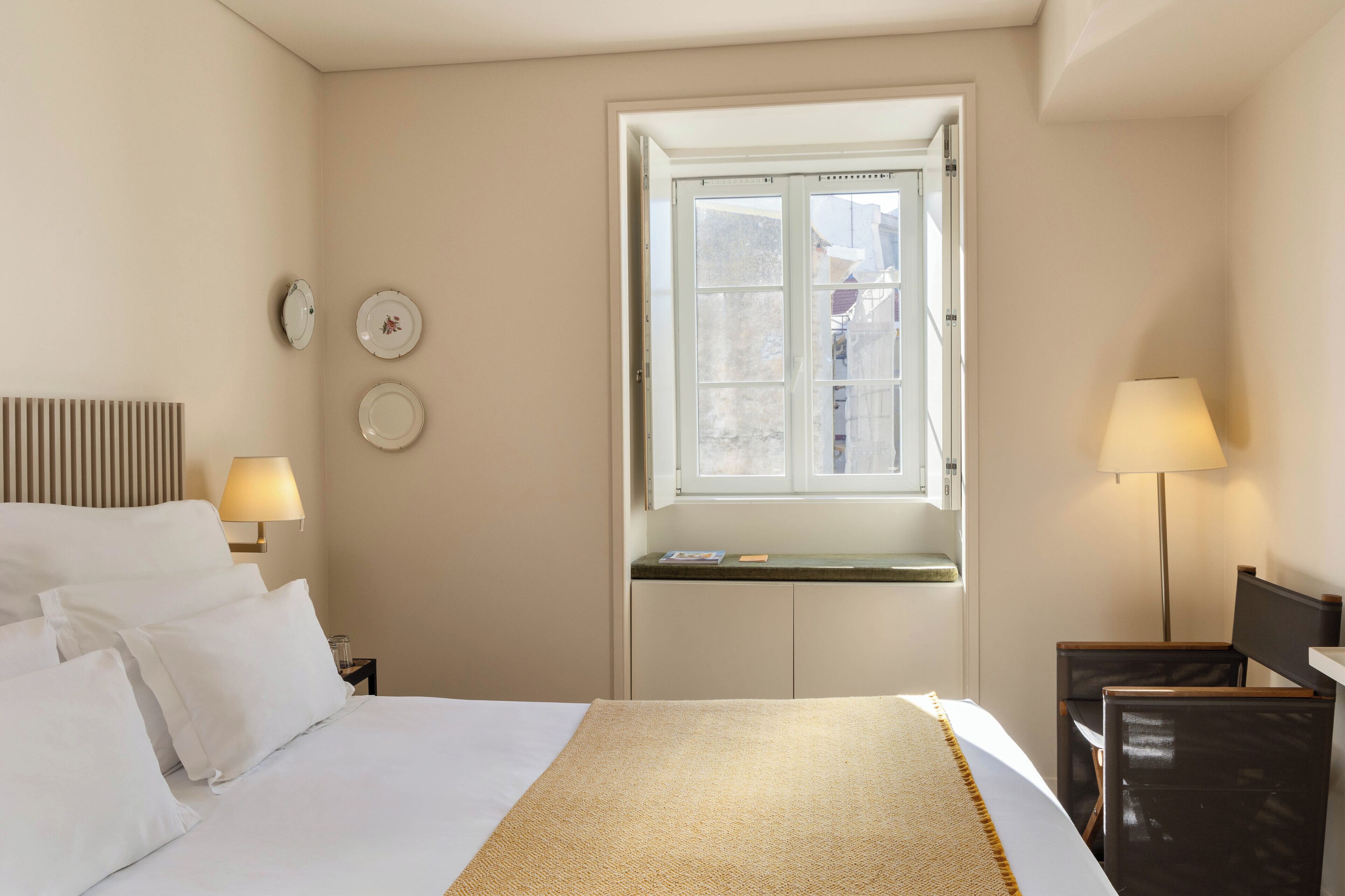 A hotel room with a bed, two bedside tables with lamps, a window with a view, and a wall-mounted shelf with a book.