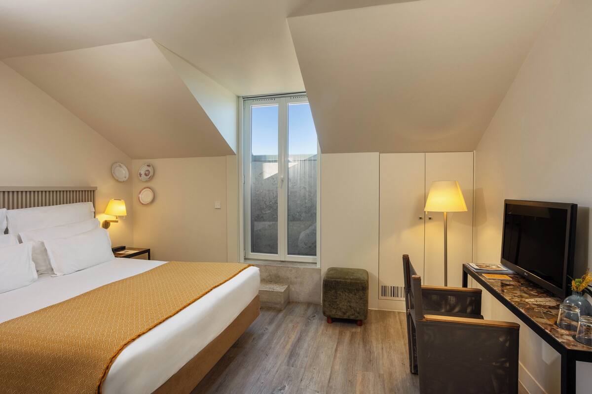 A hotel room with a bed, a desk with a television, a small stool, and a window with a view.