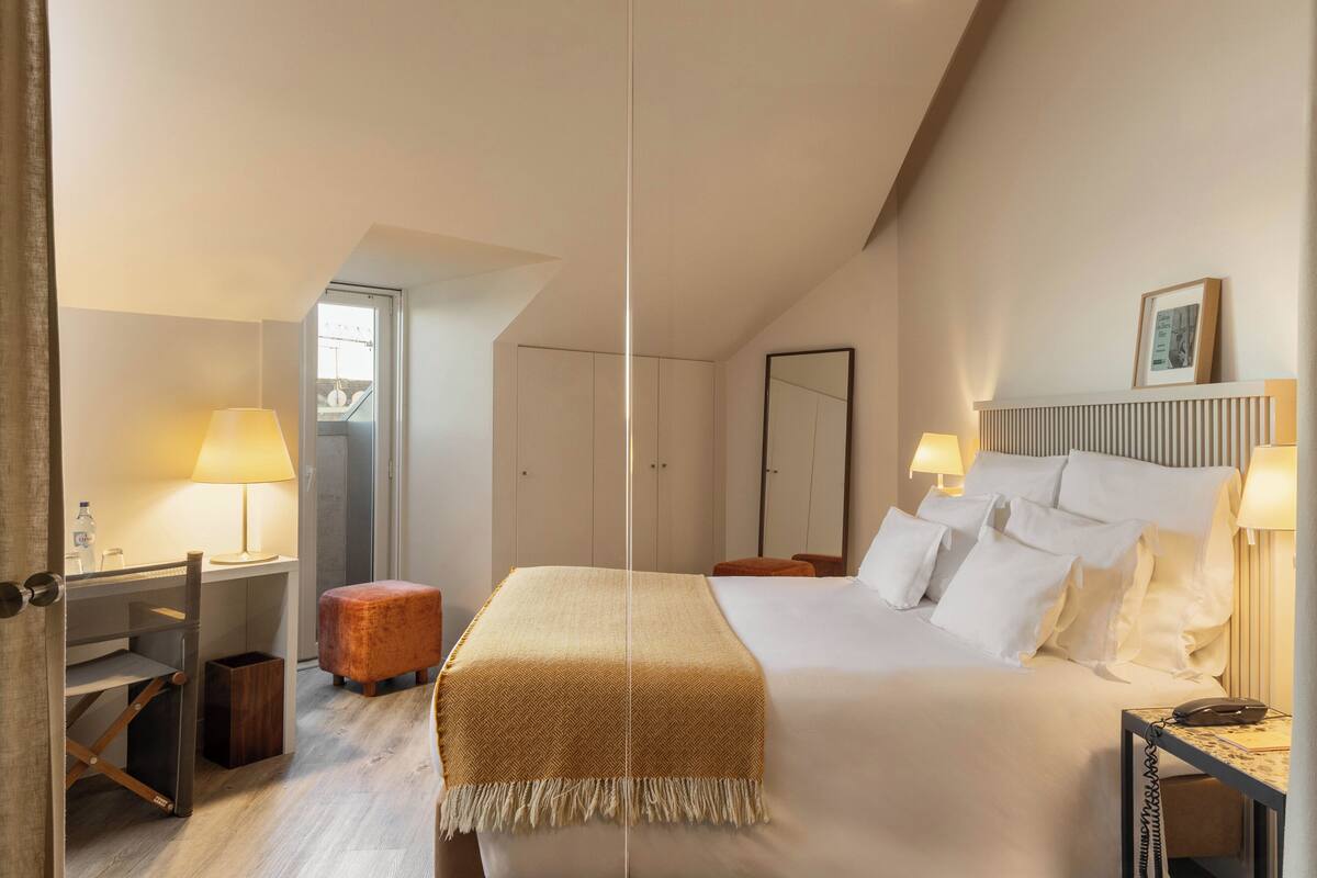 A hotel room with a bed, bedside lamps, a desk, a chair, a small stool, and a wardrobe.