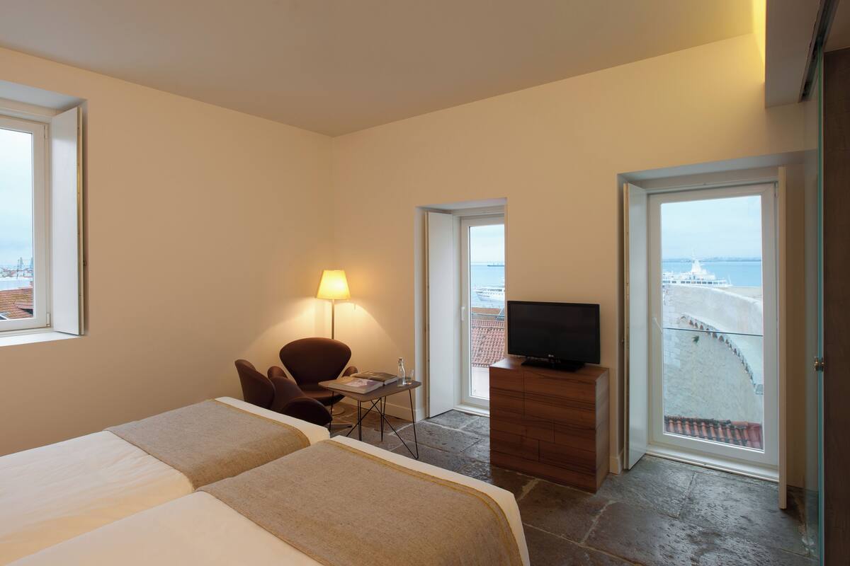 A hotel room with two beds, a desk with a lamp, a TV, and a view of the sea and buildings.