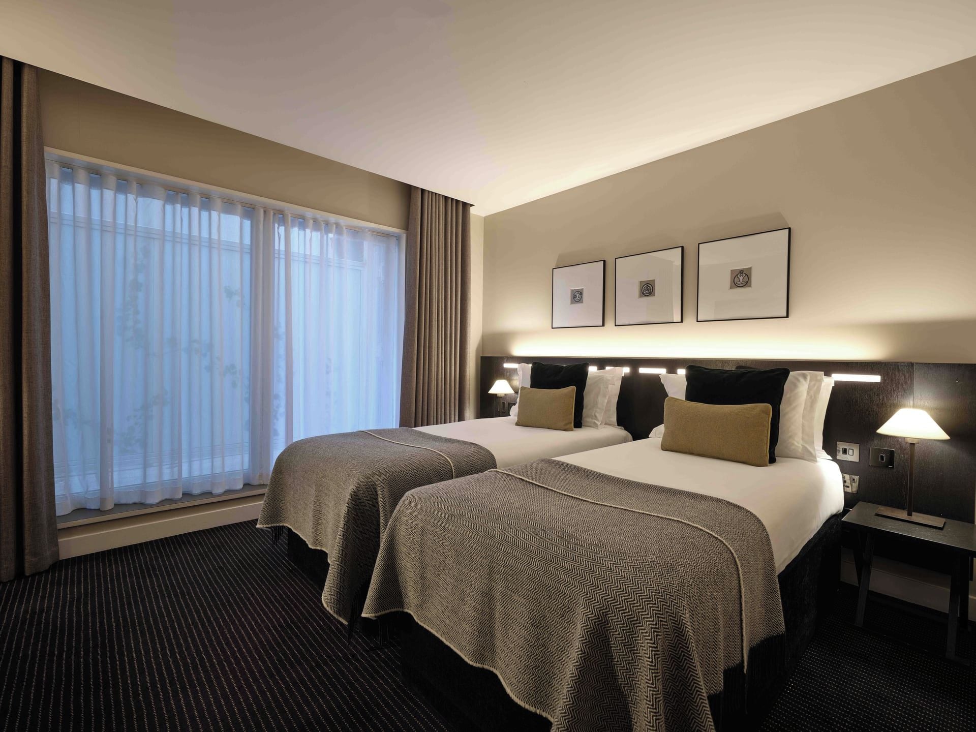 Een hotelkamer met twee bedden, een groot raam met gordijnen, een wandlamp en ingelijste foto's.
