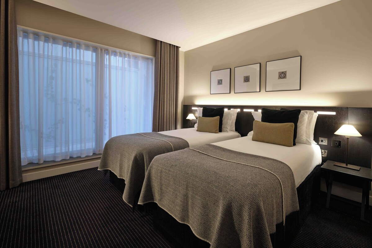 Quarto de hotel com duas camas, uma janela grande com cortinas, cabeceira com iluminação e quadros nas paredes.