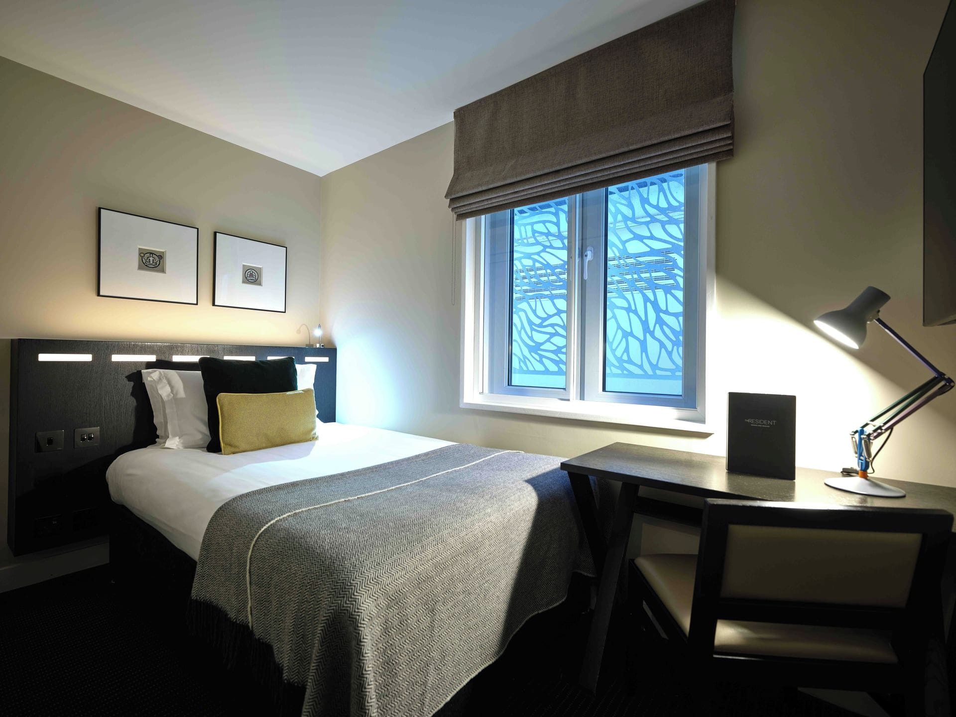 Quarto de hotel com cama, mesa de cabeceira, luminária, cadeira, escrivaninha e janela com persianas.
