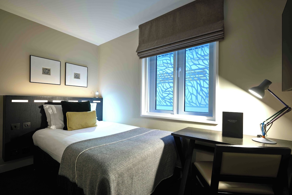 Quarto de hotel com cama, escrivaninha, cadeira, luminária e janela com persianas.