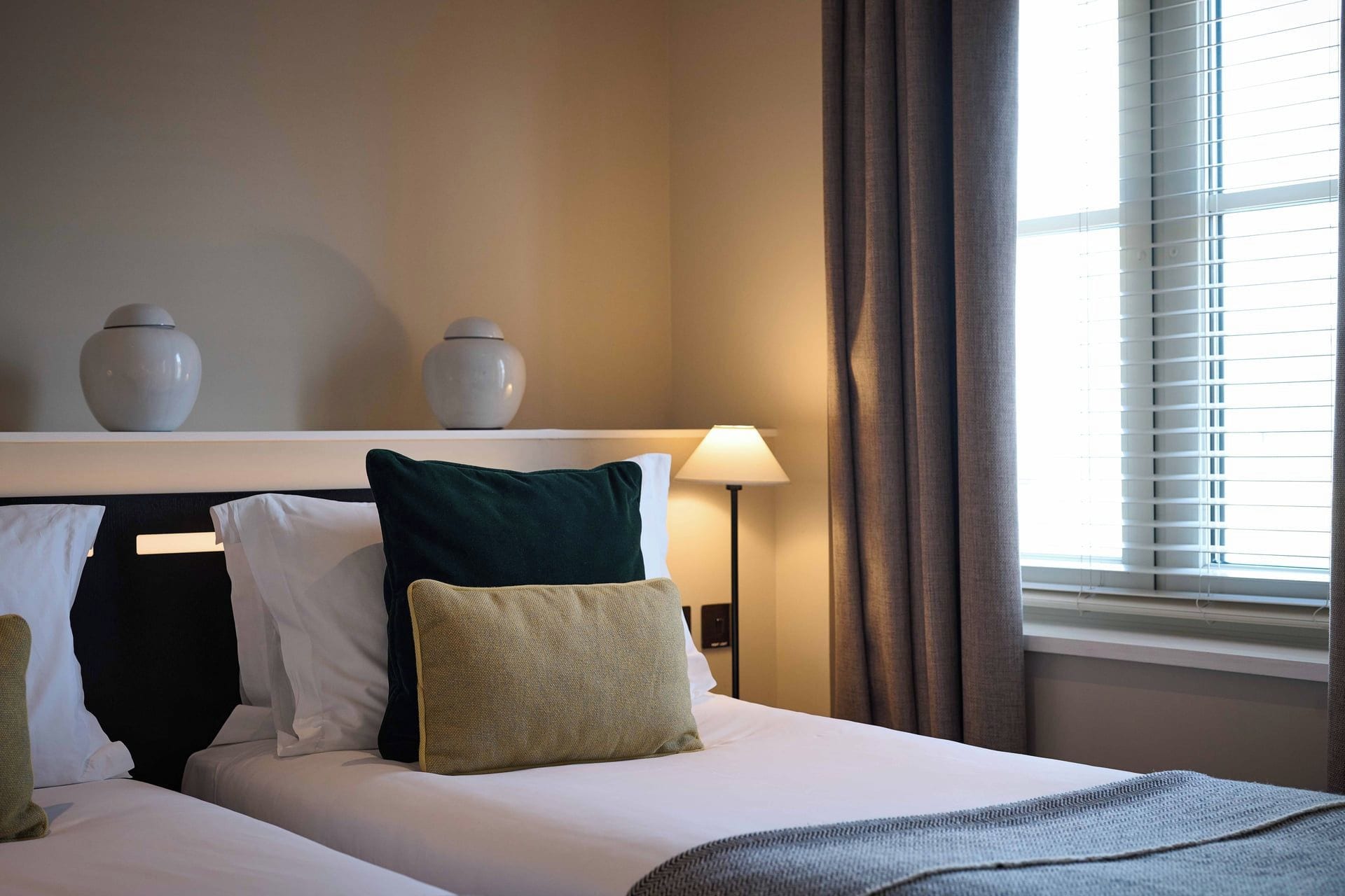 A hotel room with a bed, pillows, a lamp, and a window with blinds.