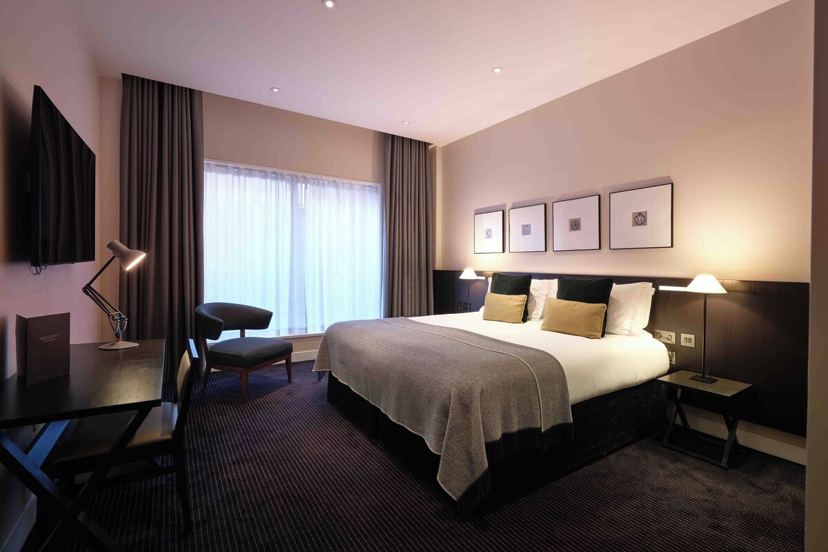 Quarto de hotel com cama, escrivaninha, cadeira, televisão e janela com cortinas.