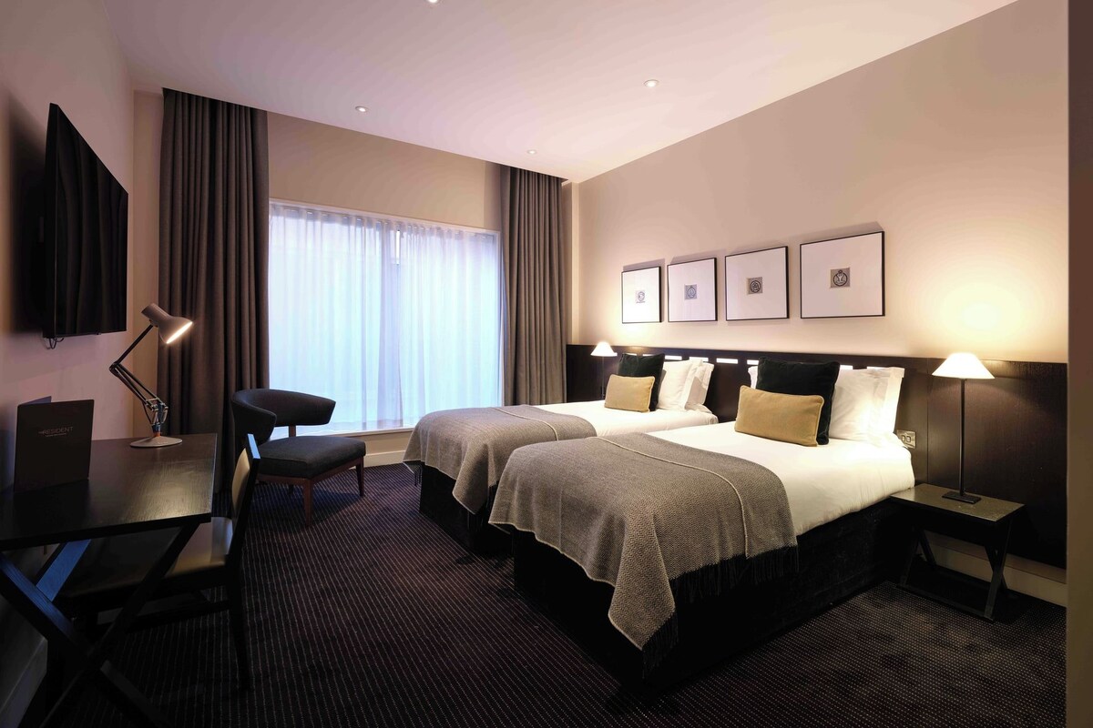 Quarto de hotel com duas camas, uma escrivaninha, uma cadeira, uma televisão e uma janela com cortinas.