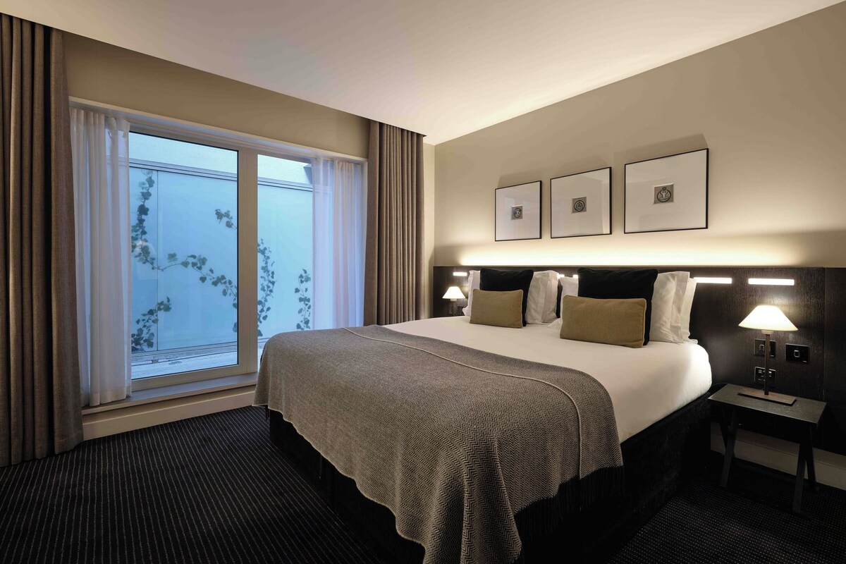 Quarto de hotel com uma cama grande, mesa de cabeceira, luminária e janela com cortinas.