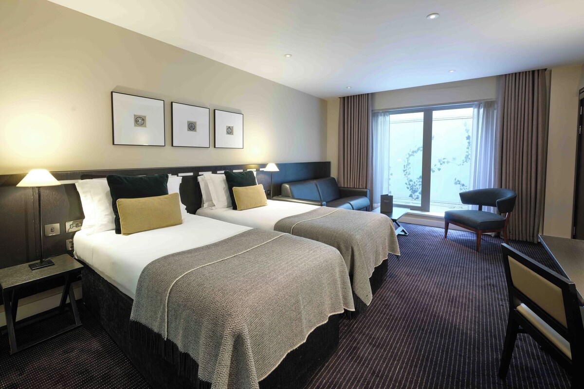 Quarto de hotel com duas camas, uma escrivaninha, uma cadeira e uma janela grande.