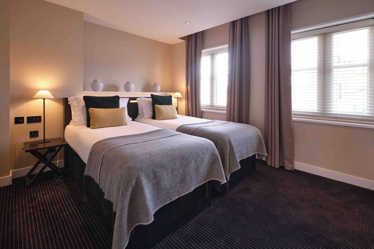 Quarto de hotel com duas camas, uma mesa de cabeceira, luminárias e uma janela com cortinas.