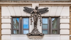 Fachada de edifício com uma estátua de figura alada segurando uma árvore, e o nome "THE RESIDENT" exibido abaixo.