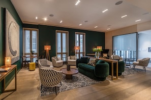 Uma sala de estar moderna com um sofá verde, duas poltronas, uma mesa de centro e um espaço de refeições com mesa e cadeiras. Há janelas amplas com persianas, um abajur e um quadro na parede.