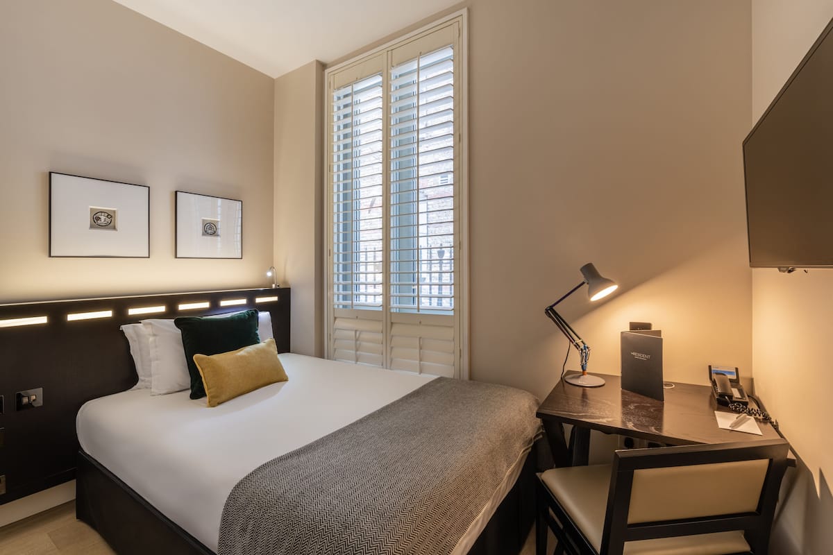 A hotel room with a bed, a desk with a lamp, a chair, and a window with blinds.