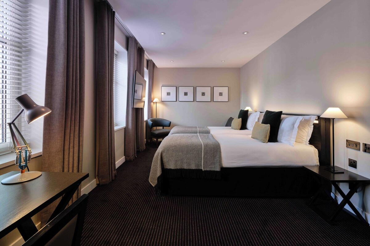 Quarto de hotel com uma cama grande, uma escrivaninha com luminária, uma cadeira e uma janela com persianas.