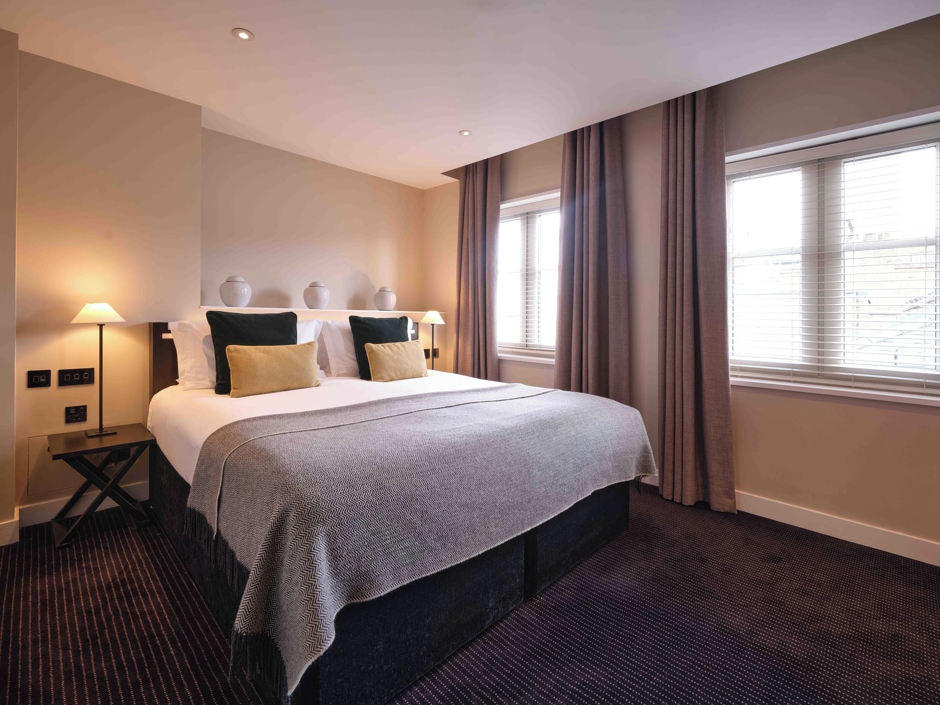A hotel room with a bed, bedside lamps, a nightstand, and a window with curtains.