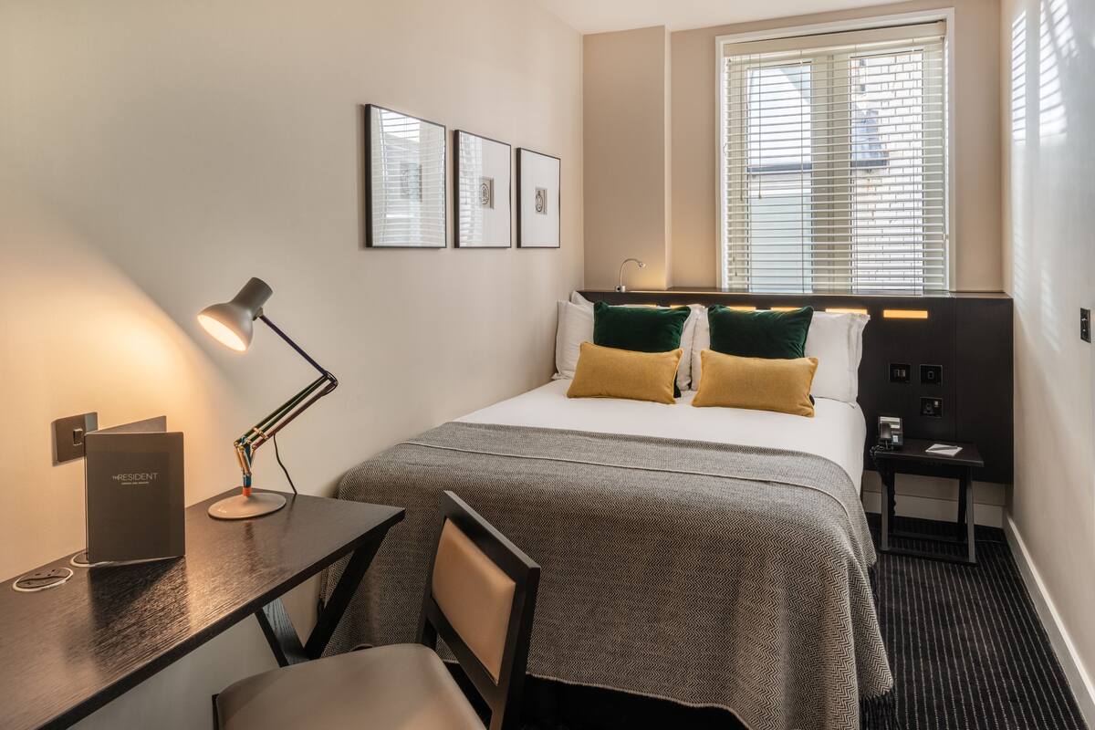A hotel room with a bed, a desk with a lamp, a chair, a nightstand, and a window with blinds.