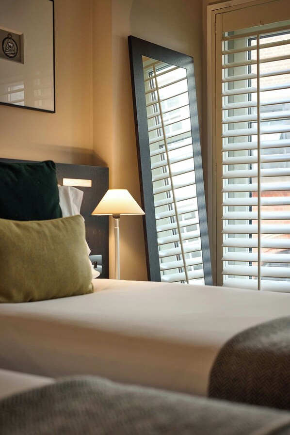 Una habitación de hotel con cama, mesita de noche, lámpara y ventana con persianas.