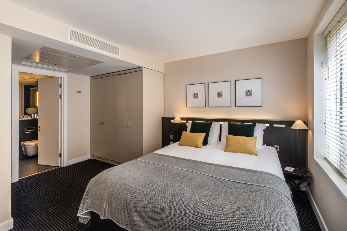 Quarto de hotel com cama, mesas de cabeceira, janela com persianas, porta para um banheiro e cabeceira com luzes fixadas na parede.