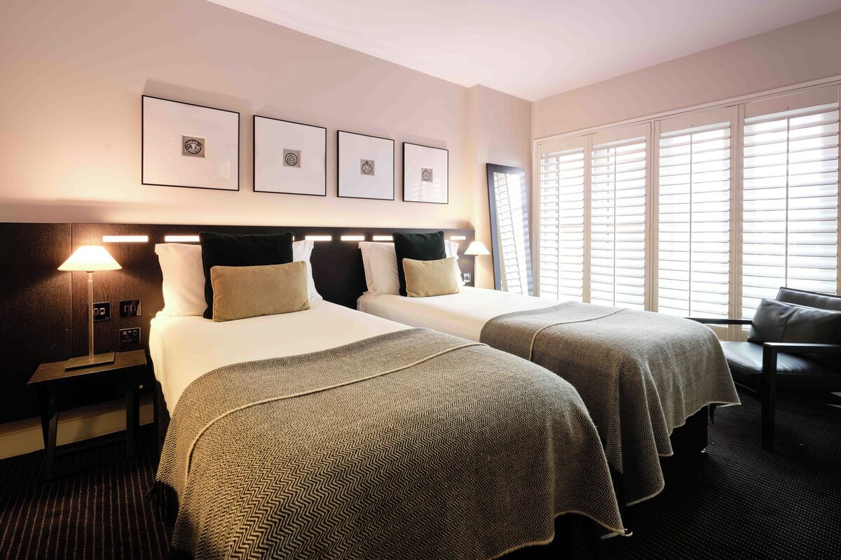 Quarto de hotel com duas camas, uma mesa de cabeceira, abajur e janela com persianas.