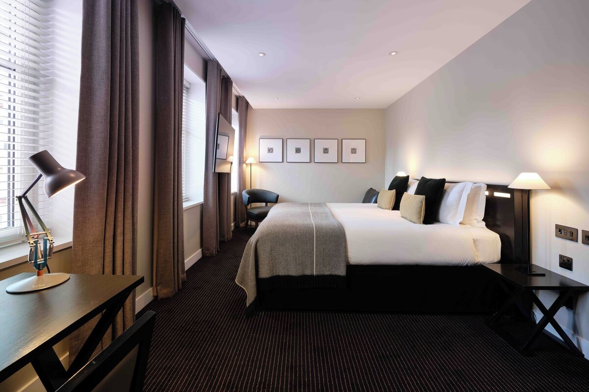 Quarto de hotel com cama, mesas de cabeceira, escrivaninha, cadeira, janela com cortinas e parede com quadros.