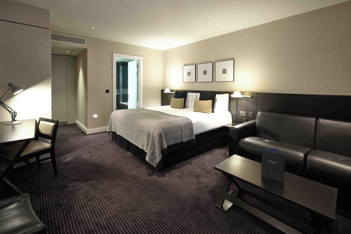 Quarto de hotel com cama, escrivaninha, cadeira, sofá e mesa pequena.
