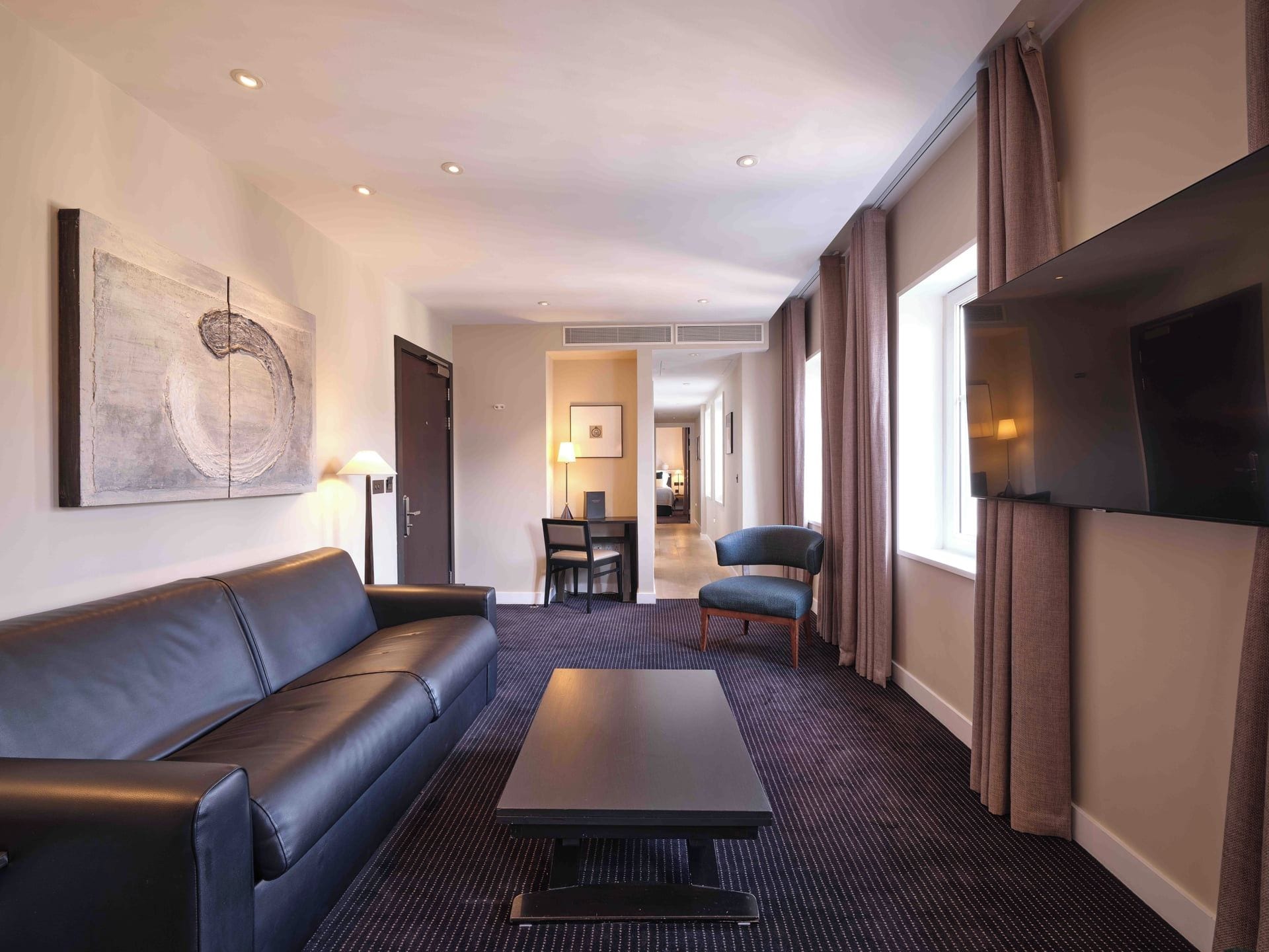 Habitación de hotel con un sofá de cuero negro, una mesita de centro, un televisor montado en la pared y vistas al pasillo.