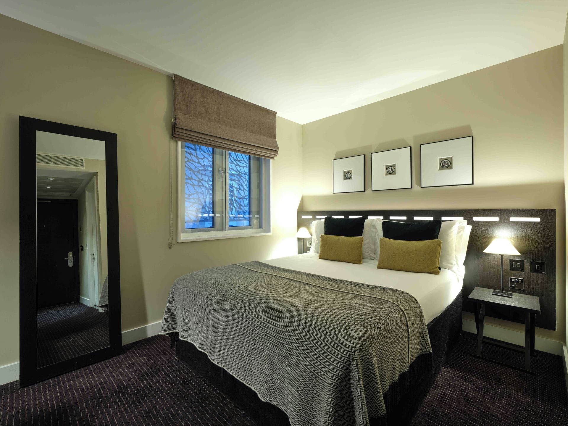 A hotel room with a bed, a nightstand, a lamp, a window with a view, and a door.