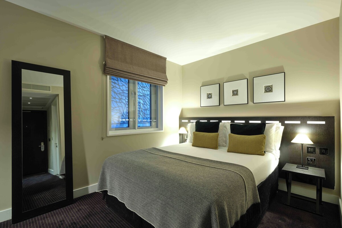 Quarto de hotel com cama, mesa de cabeceira, luminária, janela com vista e porta.