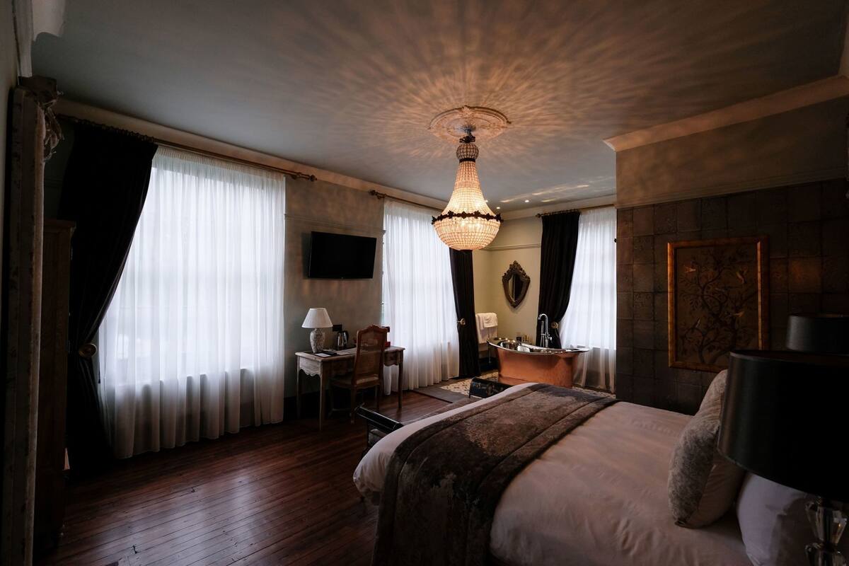 A hotel room with a large bed, a desk with a lamp, a bathtub, and a chandelier.