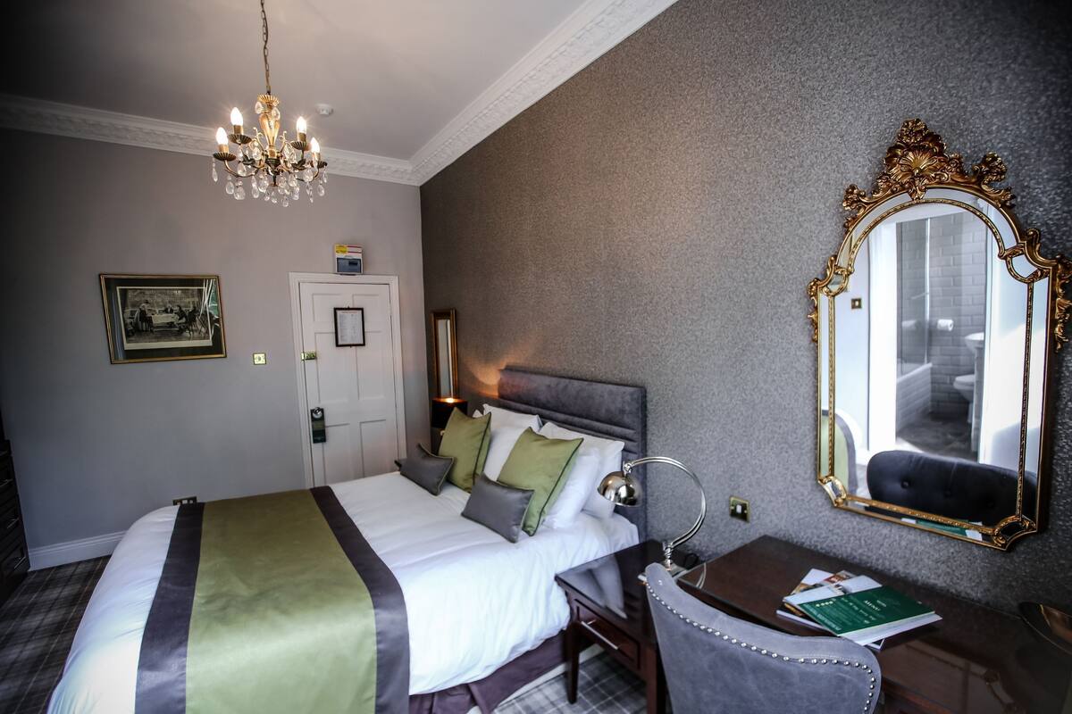 A hotel room with a large bed, a desk with a chair, a mirror, and a chandelier.