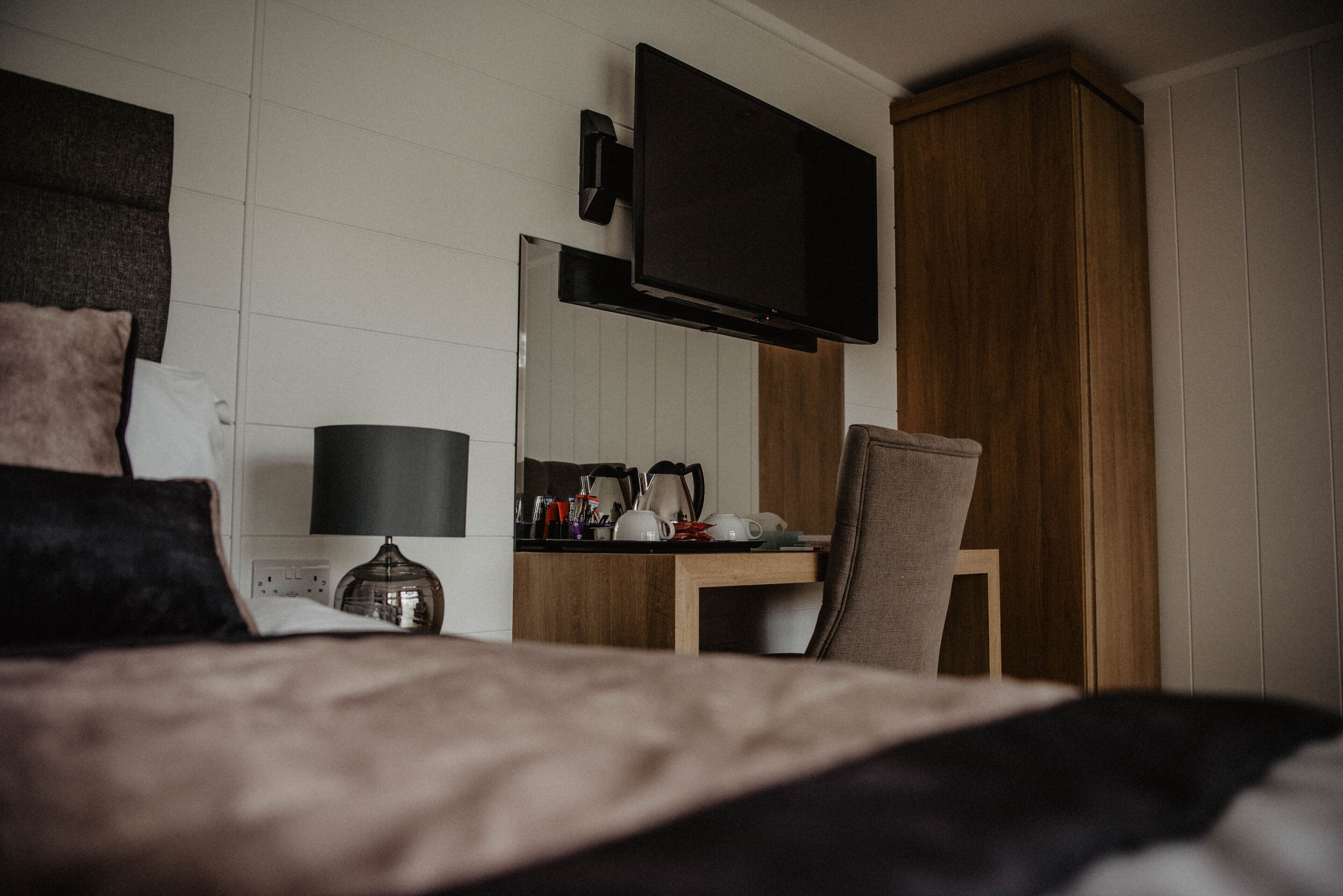Una habitación de hotel con una cama, un televisor montado en la pared, un armario de madera y una mesita con lámpara y hervidor.