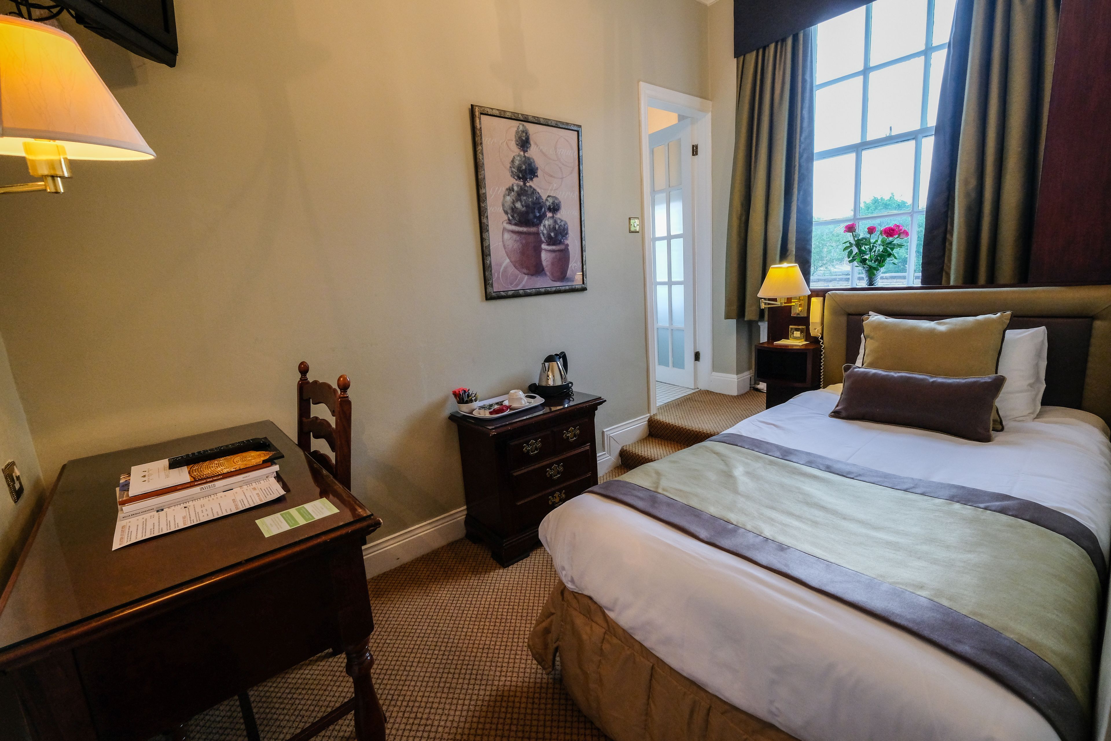 A hotel room with a bed, a desk with papers, a nightstand with a lamp, and a window with curtains.