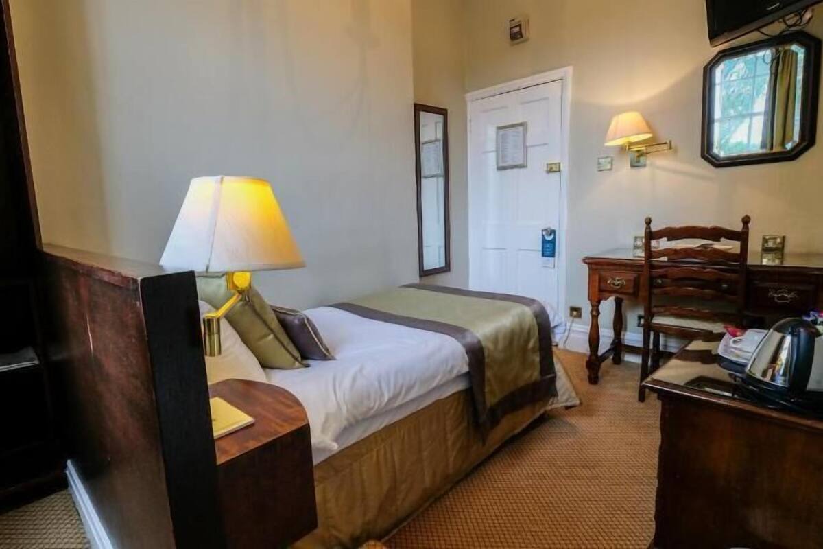 A hotel room with a bed, bedside table, lamp, desk, chair, and a small window.