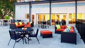 An outdoor seating area with wicker furniture and orange cushions, a round table, and a view of the building's interior through large windows. - Aloft by Marriott Tulsa Downtown (Tulsa)