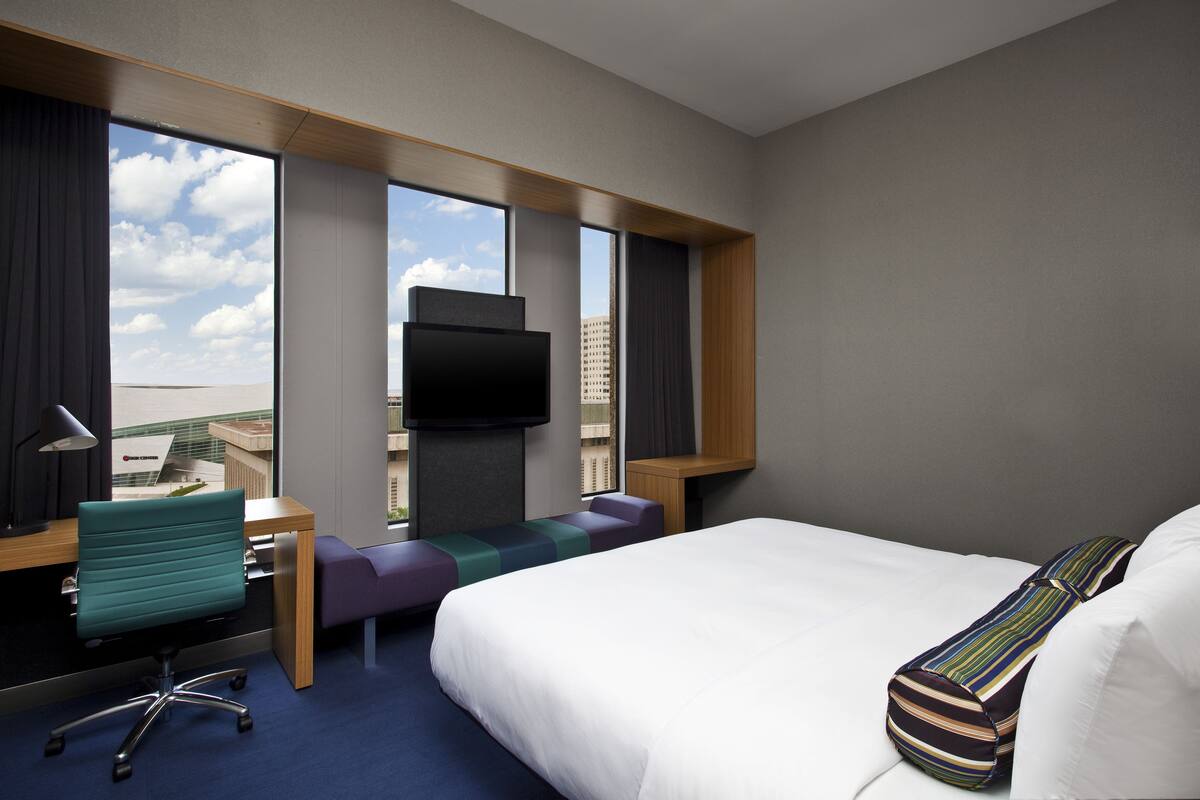 Ett hotellrum med en stor säng, ett skrivbord med en stol, en tv och utsikt över stadsbilden genom stora fönster.