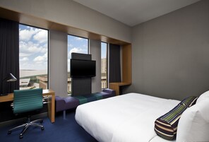 Habitación de hotel con una cama grande, un escritorio con silla, televisor y vistas de la ciudad a través de amplios ventanales.