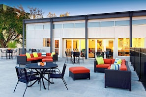 Un área de estar al aire libre con mobiliario de mimbre y una mesa redonda, frente a un edificio con grandes ventanales y diseño moderno.