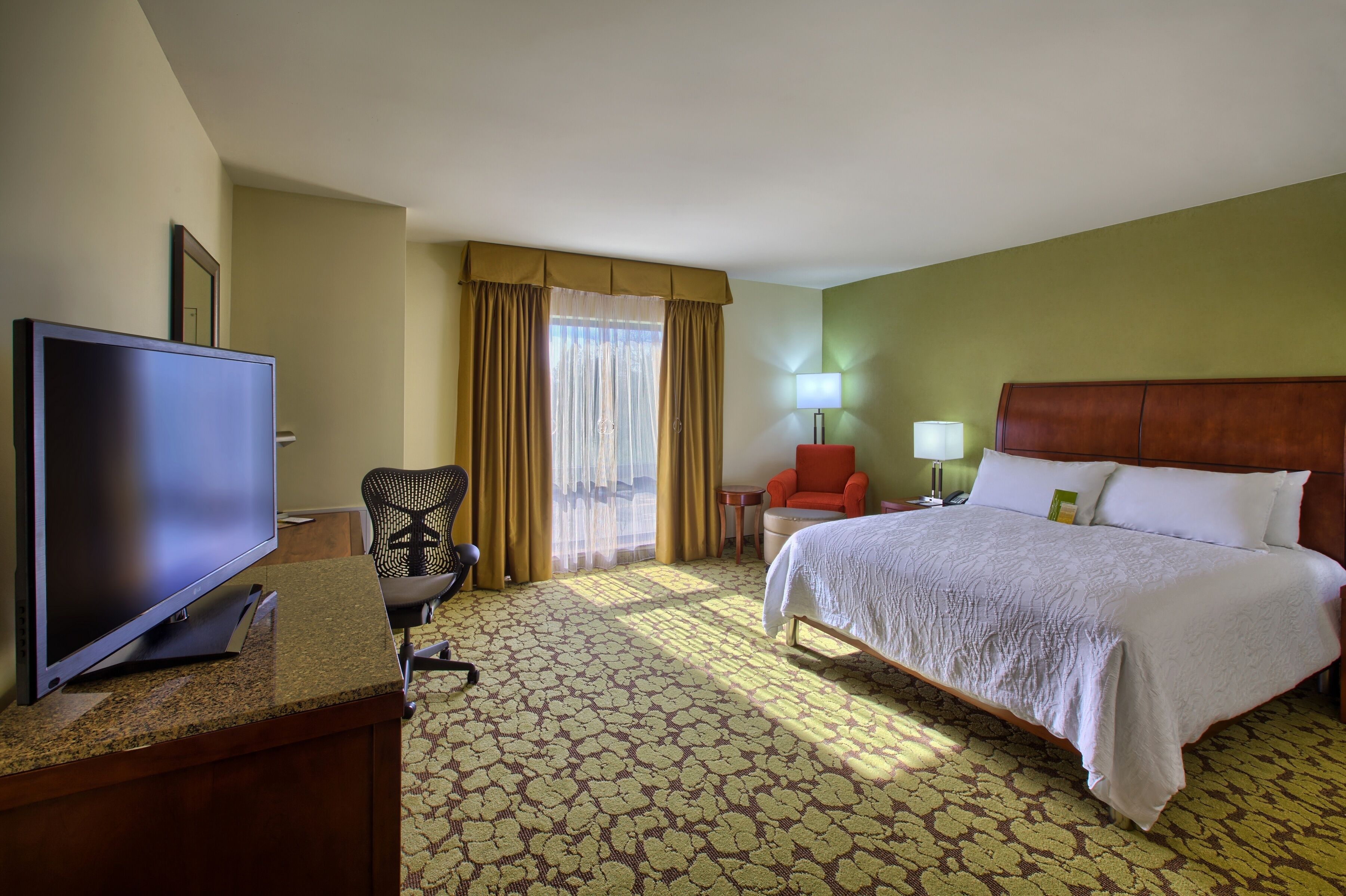 A hotel room with a bed, a desk with a television, a chair, a nightstand, and a lamp.