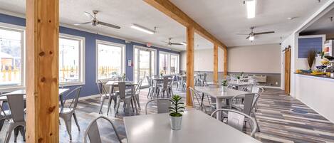 A spacious indoor dining area with multiple tables and chairs, ceiling fans, and large windows.