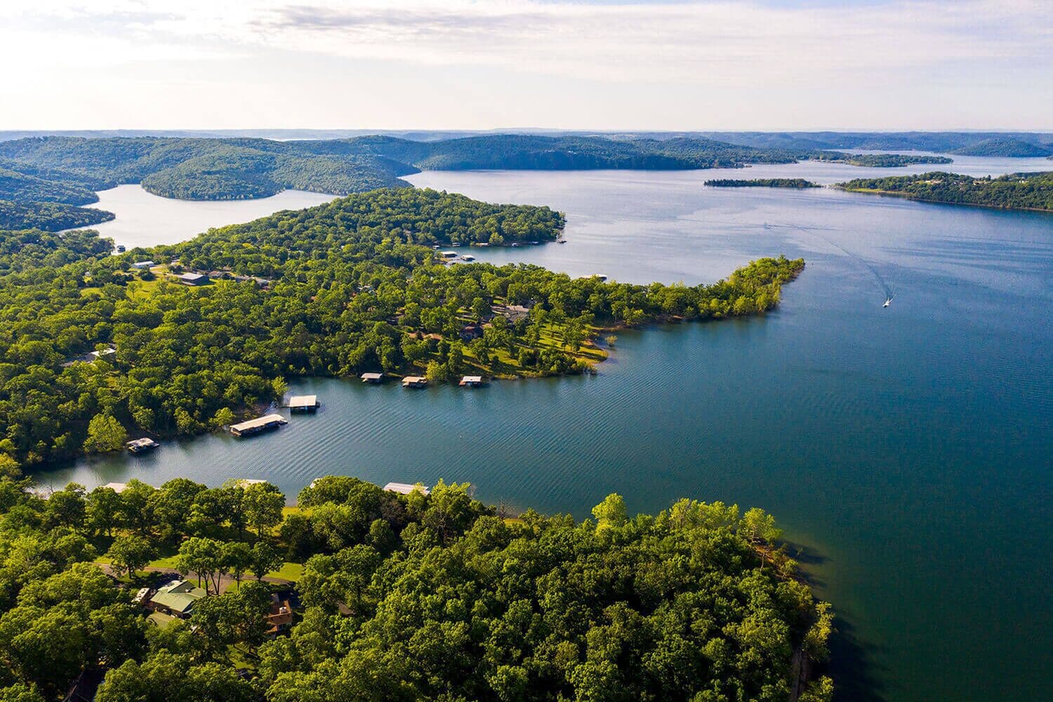 An aerial view of a lakeside property with multiple docks and boats, surrounded by dense forest and hills in the background.