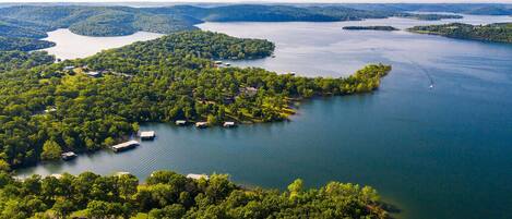 An aerial view of a lakeside property with multiple docks and boats, surrounded by dense forest and hills in the background.