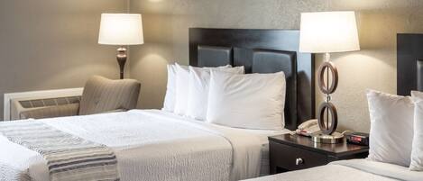 Una habitación de hotel con dos camas, cada una con sábanas blancas y mantas a rayas. Hay dos mesitas de noche con lámparas y un reloj.