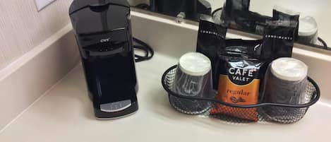 Una cafetera, un cesto con cápsulas de café y condimentos sobre una encimera.