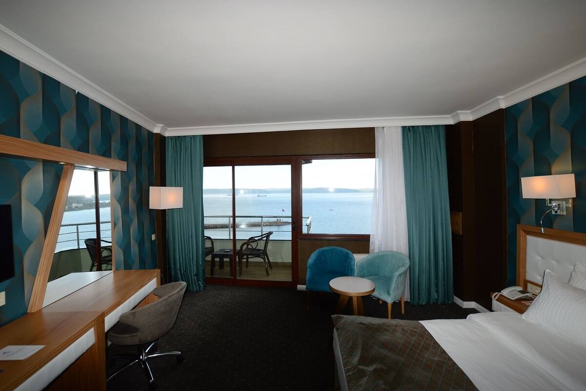 Habitación de hotel con cama, escritorio, silla, mesita y vista al mar a través de un ventanal grande.